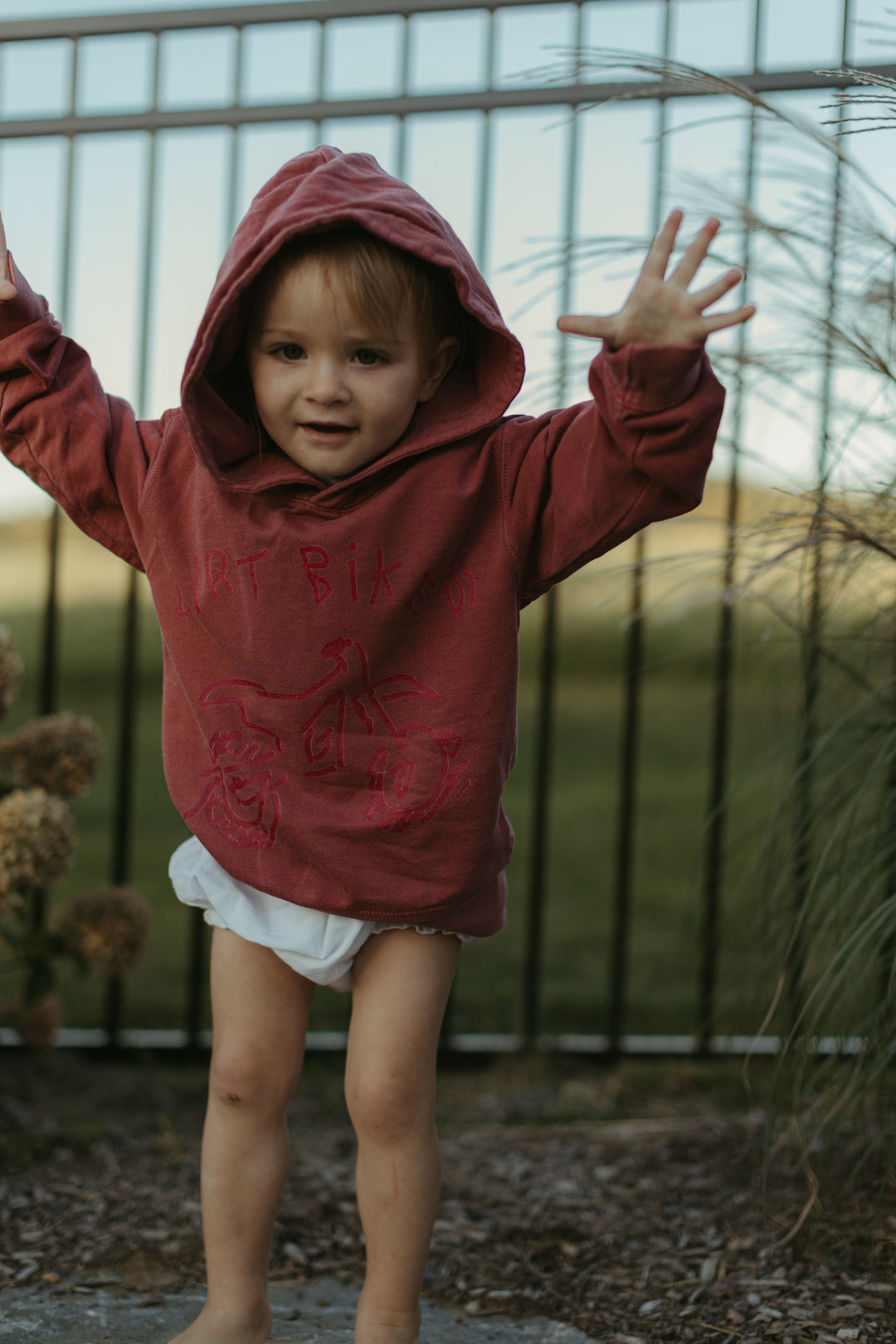 Child wearing a red hoodie with arms outstretched in front of a metal fence.