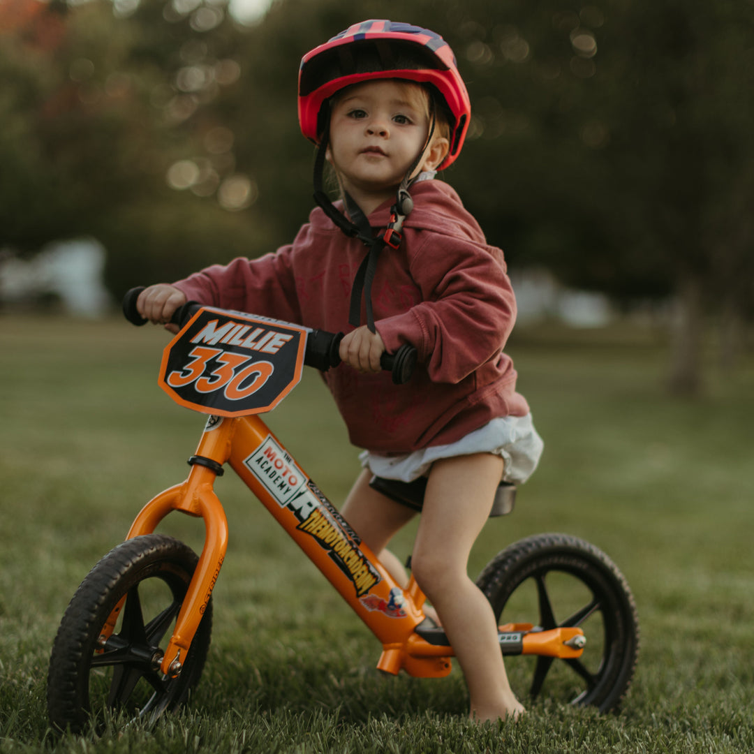 Child riding an orange balance bike in a grassy field with trees in the background