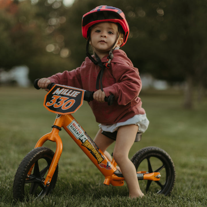 Child riding an orange balance bike in a grassy field with trees in the background